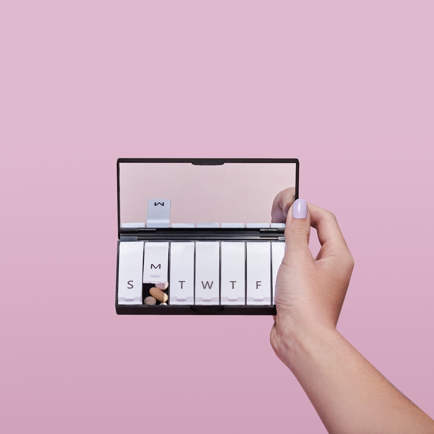 A hand holding an open Tabuu 7 day case with a mirror in the lid, on a pink backdrop